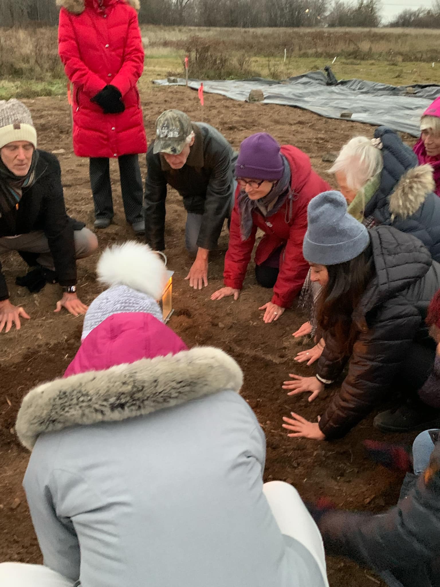 Plusieurs adultes agenouillés en extérieur, leurs mains touchant la terre sombre d'un champ en hiver.