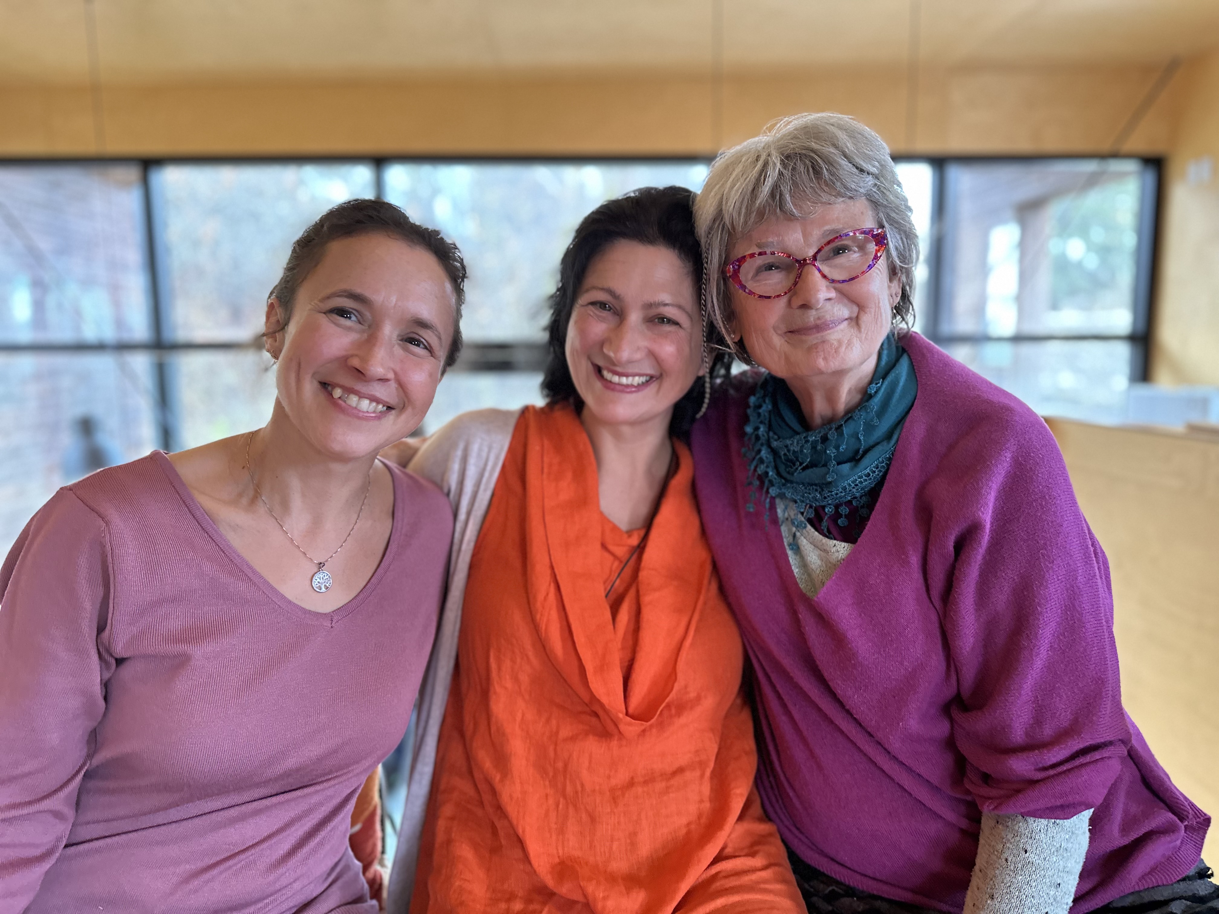 Portrait de trois femmes souriantes et joyeuses, lunettes rouges et collier arbre de vie, en intérieur.