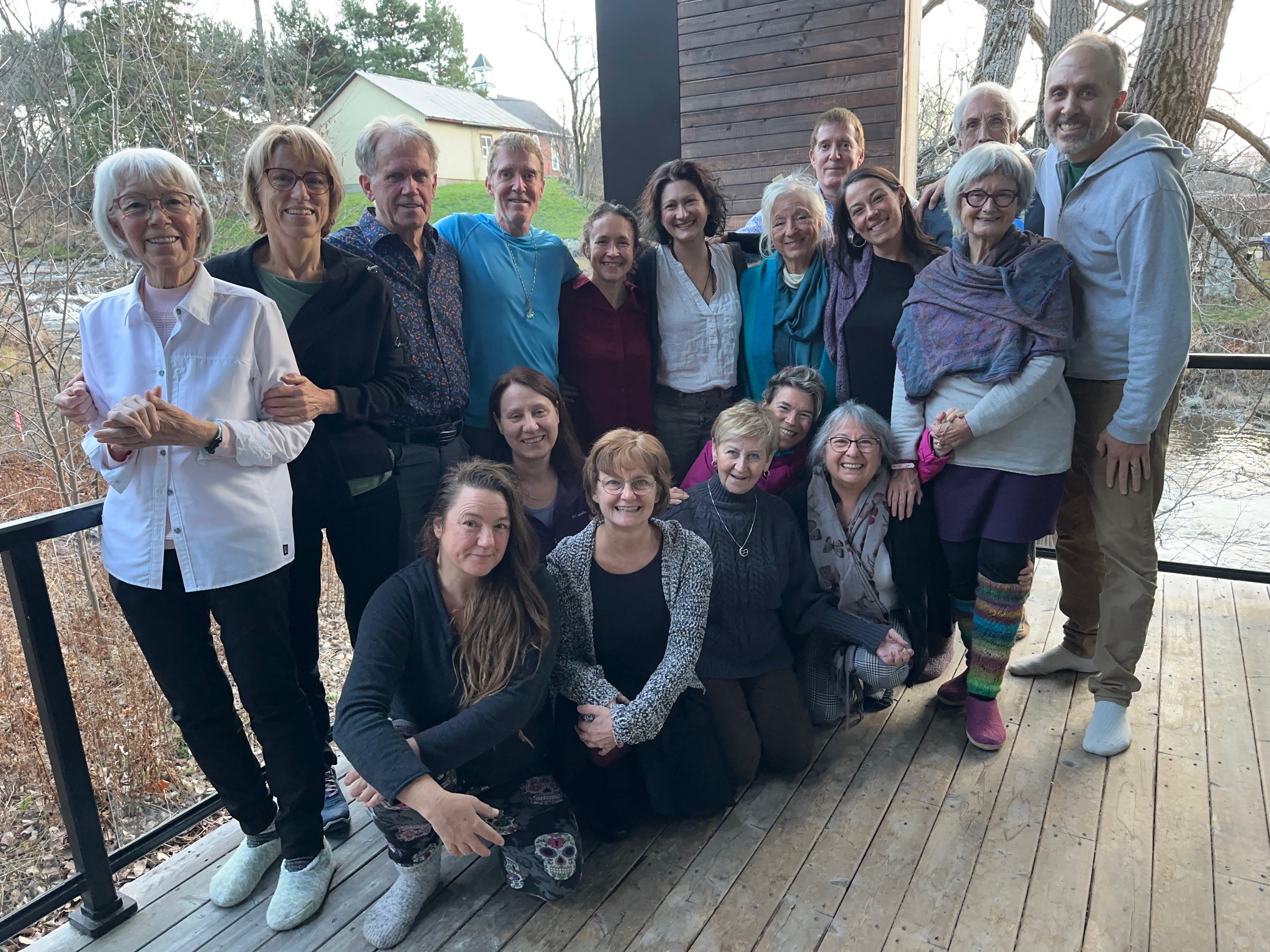 Grand groupe joyeux d'hommes et femmes souriants posant sur une terrasse en bois près d'une rivière.