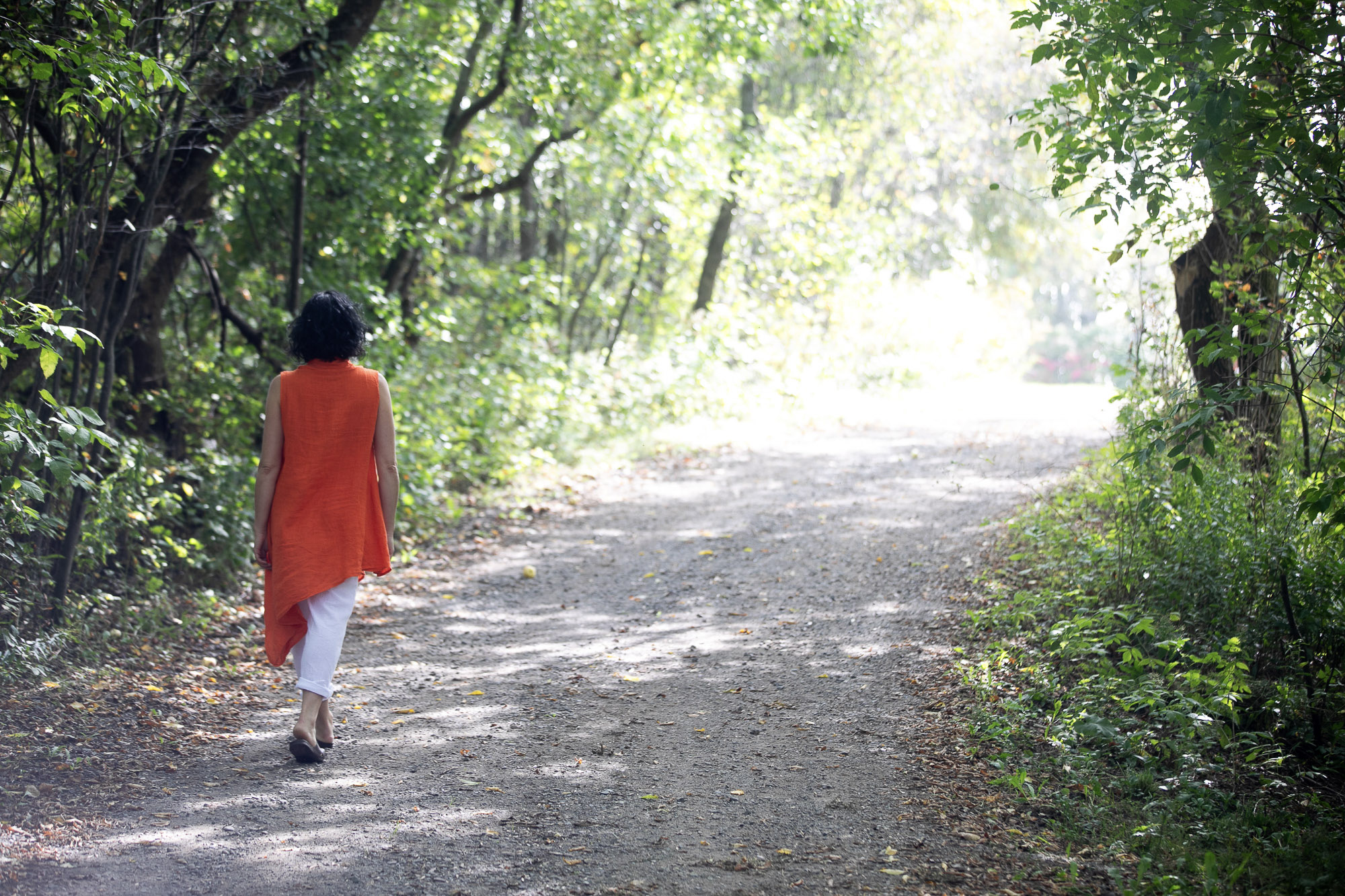 Christine Dion marche sur un chemin boisé ensoleillé, vêtue d'un haut orange et d'un pantalon blanc.