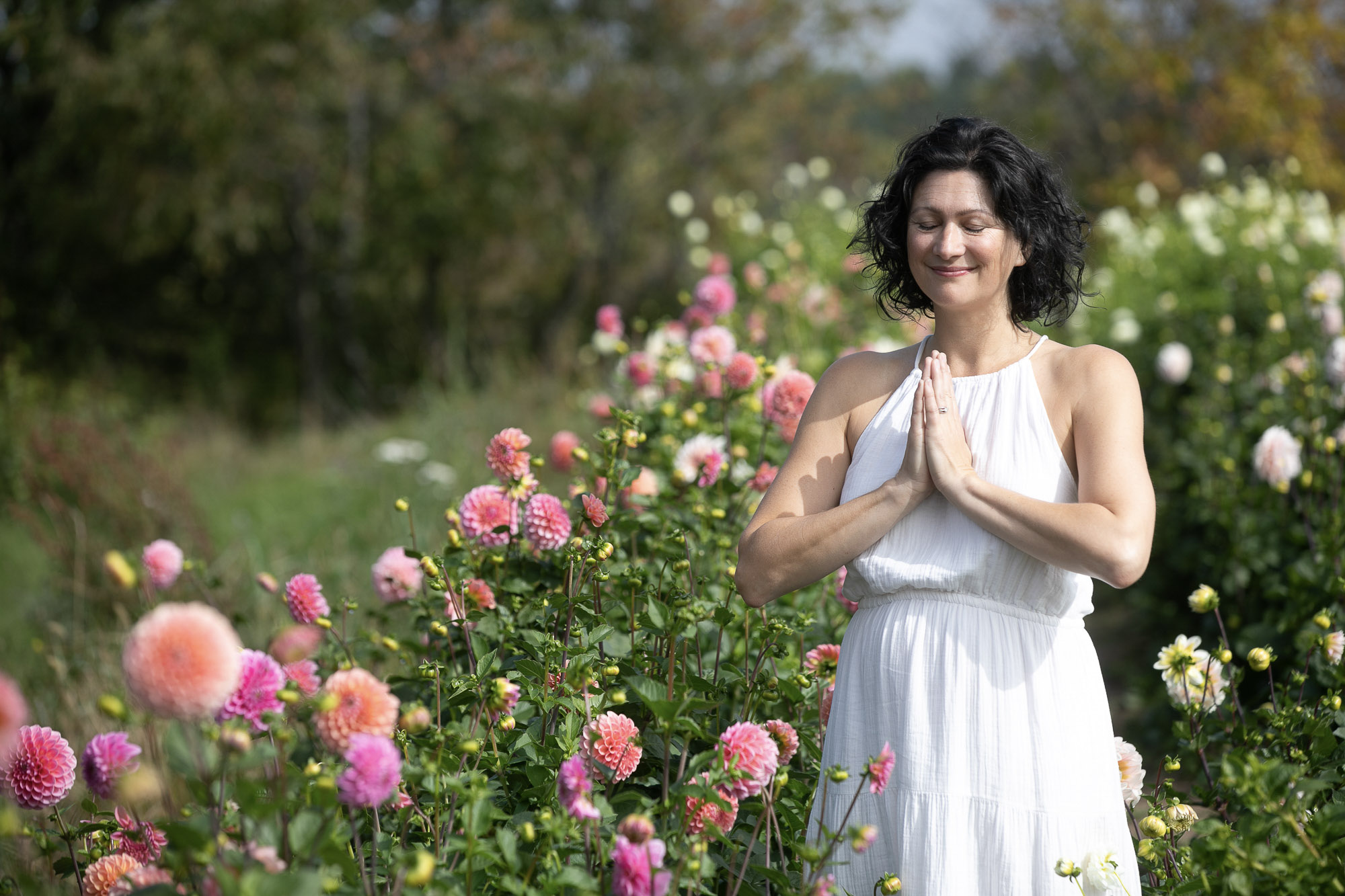 Christine Dion méditant au milieu d'un champ de dahlias colorés, les mains jointes et les yeux clos, vêtue d'une robe blanche.