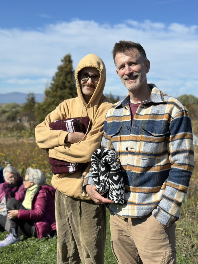 Deux hommes souriants avec des couvertures colorées, dont une à motifs noir et blanc, lors d'un rassemblement en plein air.