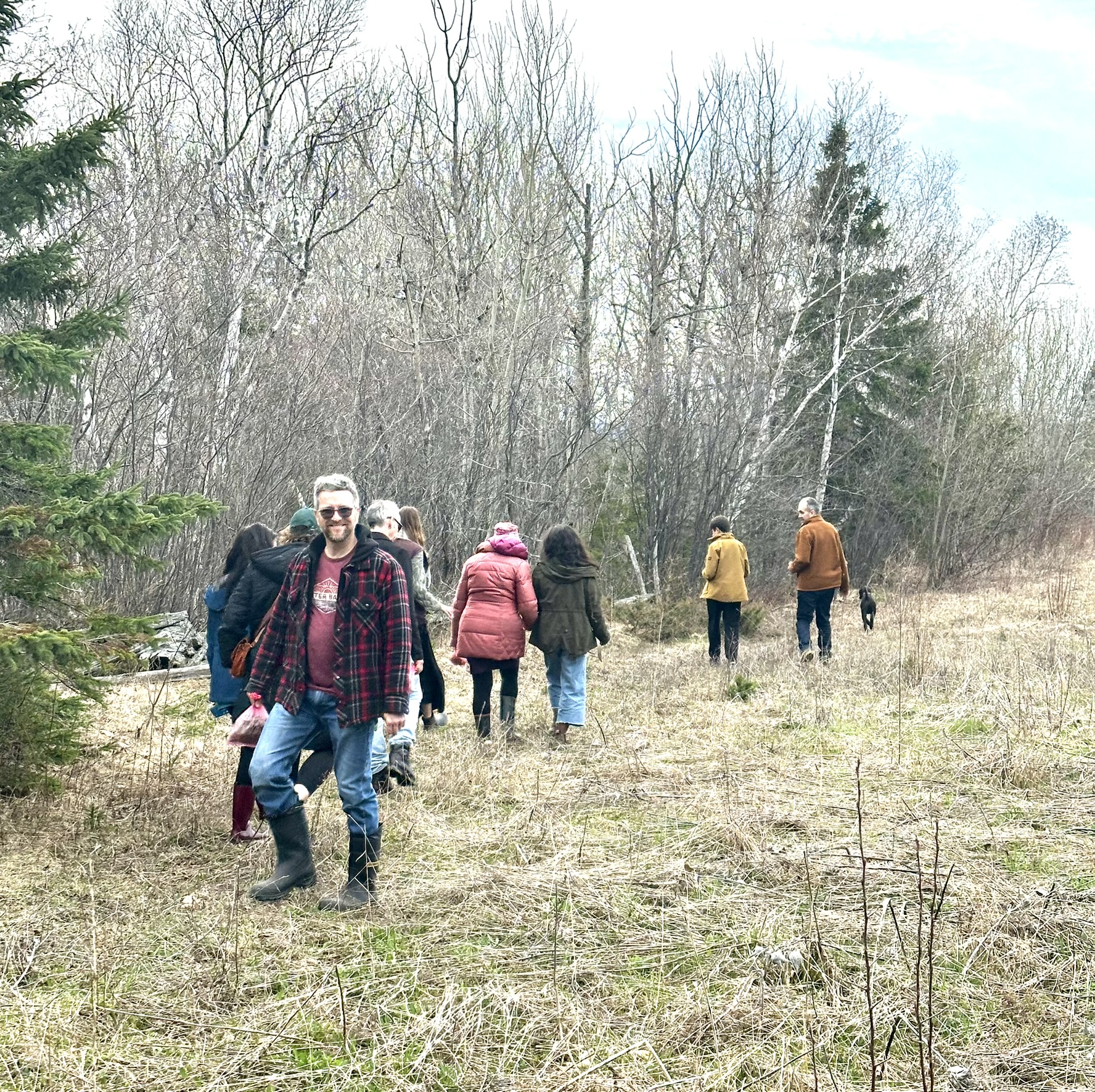 Groupe de personnes marchent sentier boisé début printemps. Homme souriant plaid premier plan, chien suit.