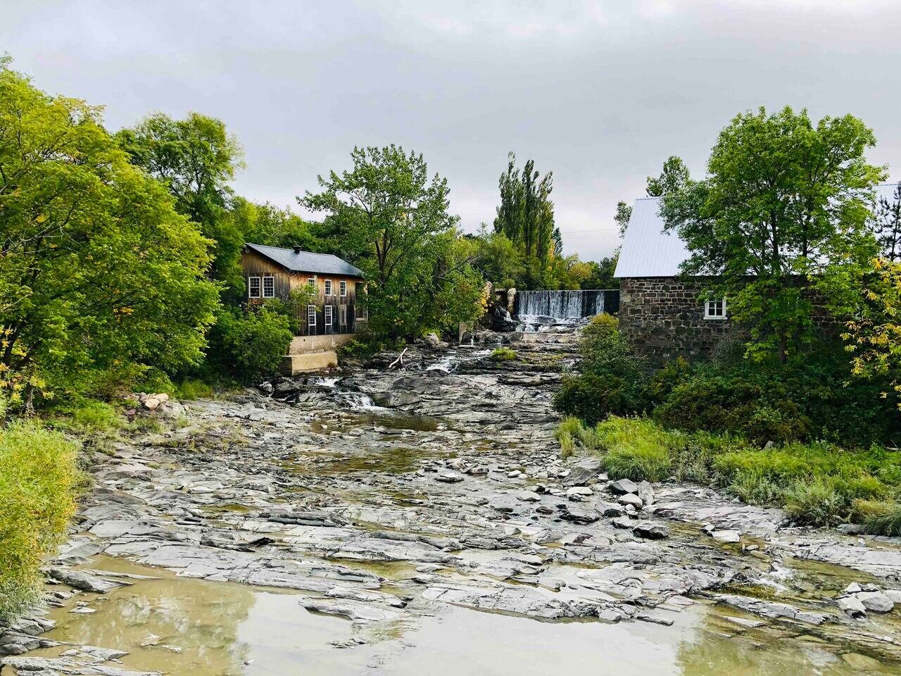 Vue d'un lit de rivière rocailleux avec peu d'eau, entouré de forêts, de bâtiments anciens et d'une petite chute d'eau.