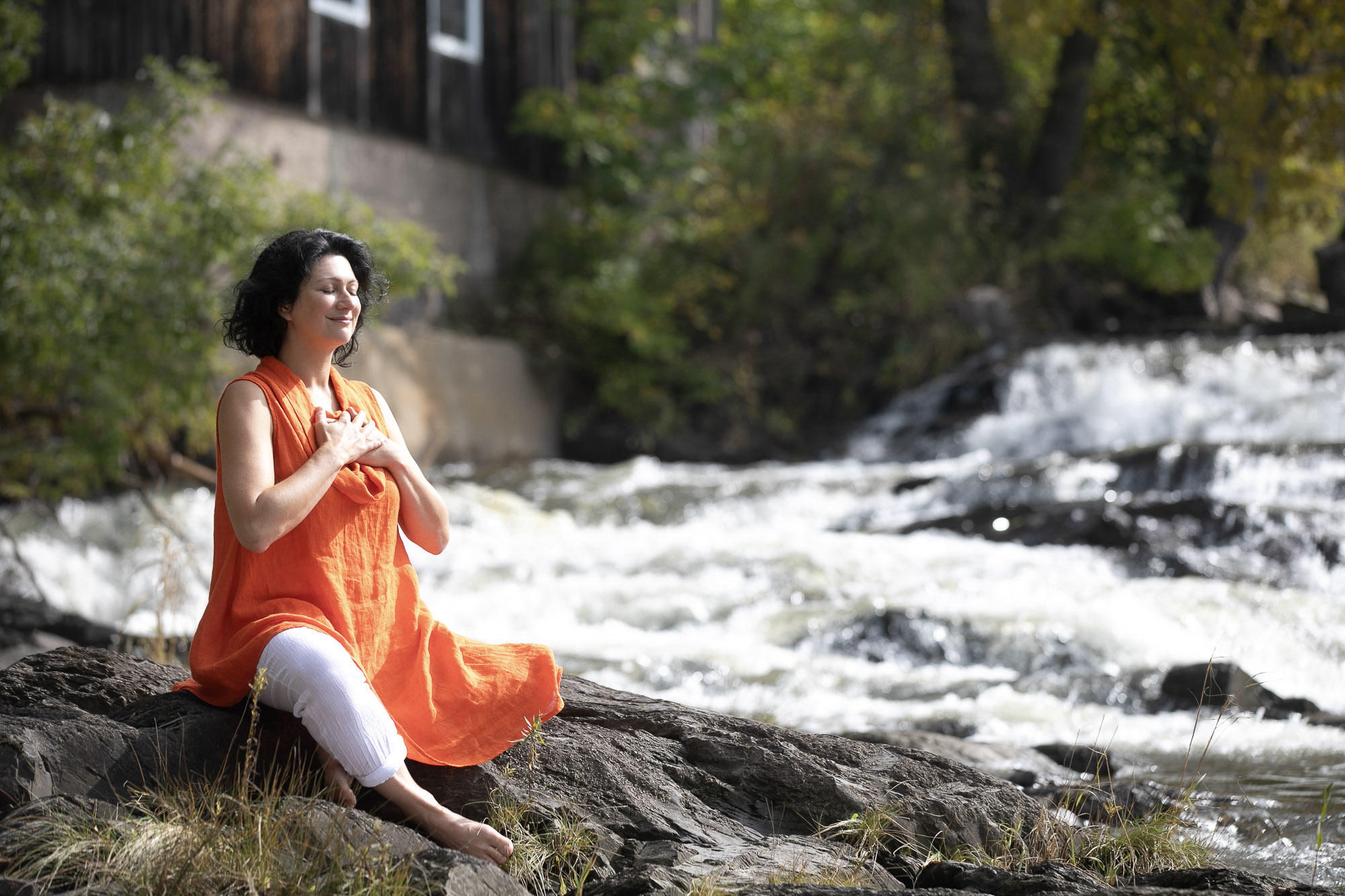 Christine Dion méditant, les mains sur le cœur, assise sur un rocher au bord d'une rivière avec des rapides.
